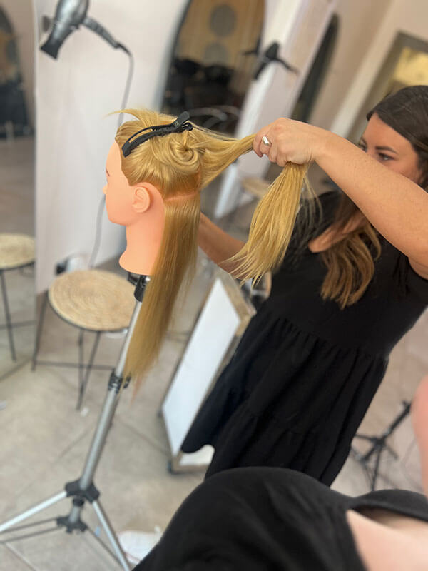 A woman practicing hair extension techniques on a training mannequin. The mannequin has long blonde hair, and the woman is sectioning and preparing to apply extensions. The setting appears to be a professional training environment, with salon equipment and tools visible in the background.