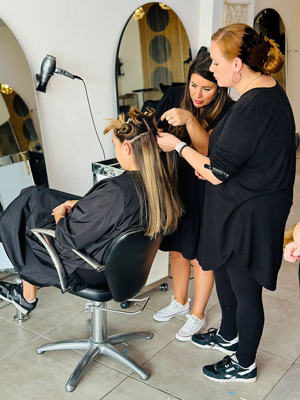 Two hairstylists work on a seated client's hair in a salon, expertly handling the Hair Extension Course by Image London. The client has partially sectioned hair and is wearing a black cape. The stylists, dressed in black, focus on their work with hair tools and brushes, while mirrors in the background reflect the scene.