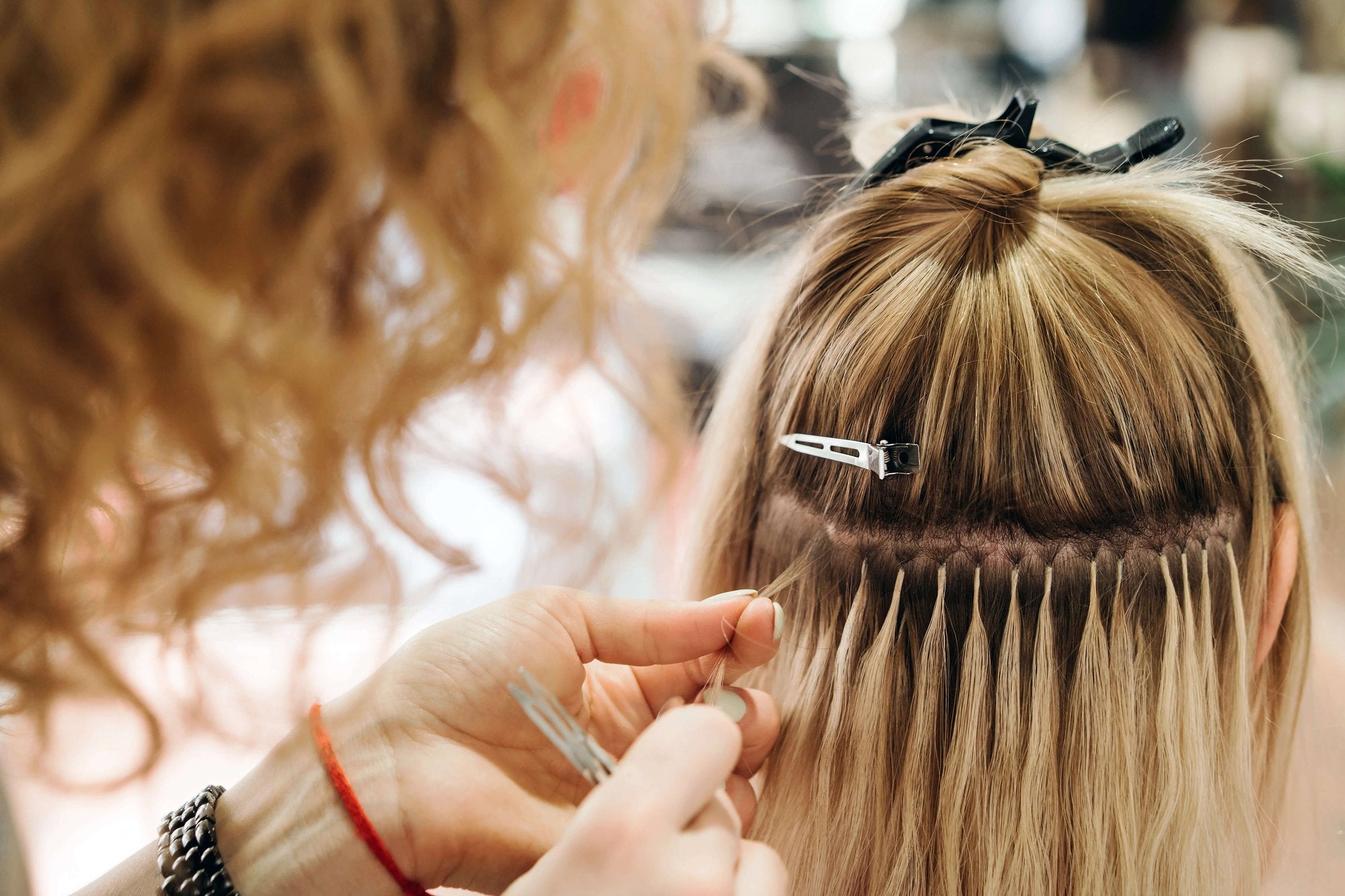 A close-up of a stylist applying hair extensions to a client's head. The stylist's hands are visible as they attach individual strands using a technique involving small clips and rings. The client has blonde hair, sectioned off and held with hair clips.