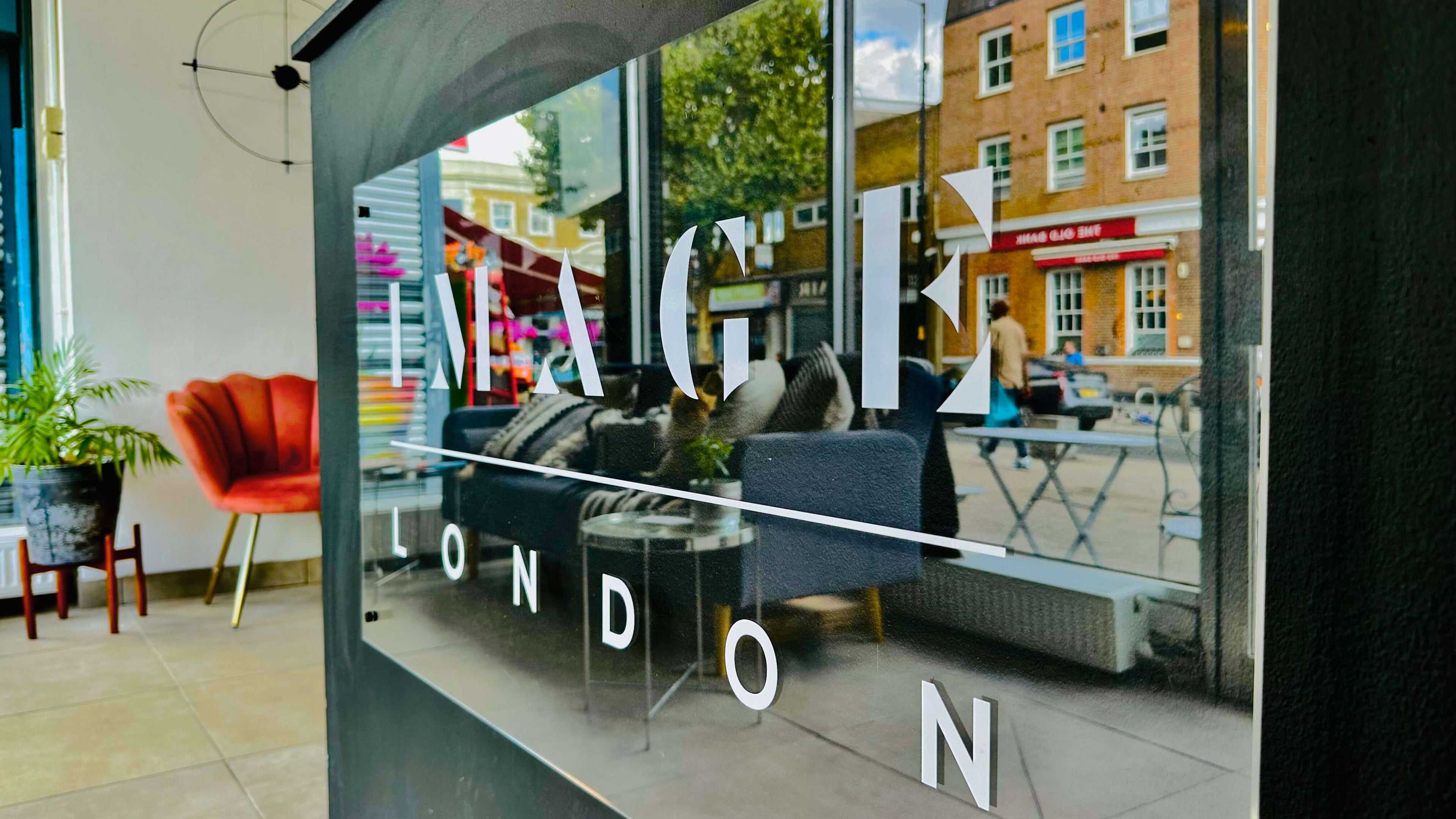 A street view of a café with the name "Image London" on the window. Inside, there's a red chair and a vibrant green plant. Outside, the reflection in the window shows the street with a pub named "The Old Bank" and other buildings across the road, subtly promoting Hair Extension Course.