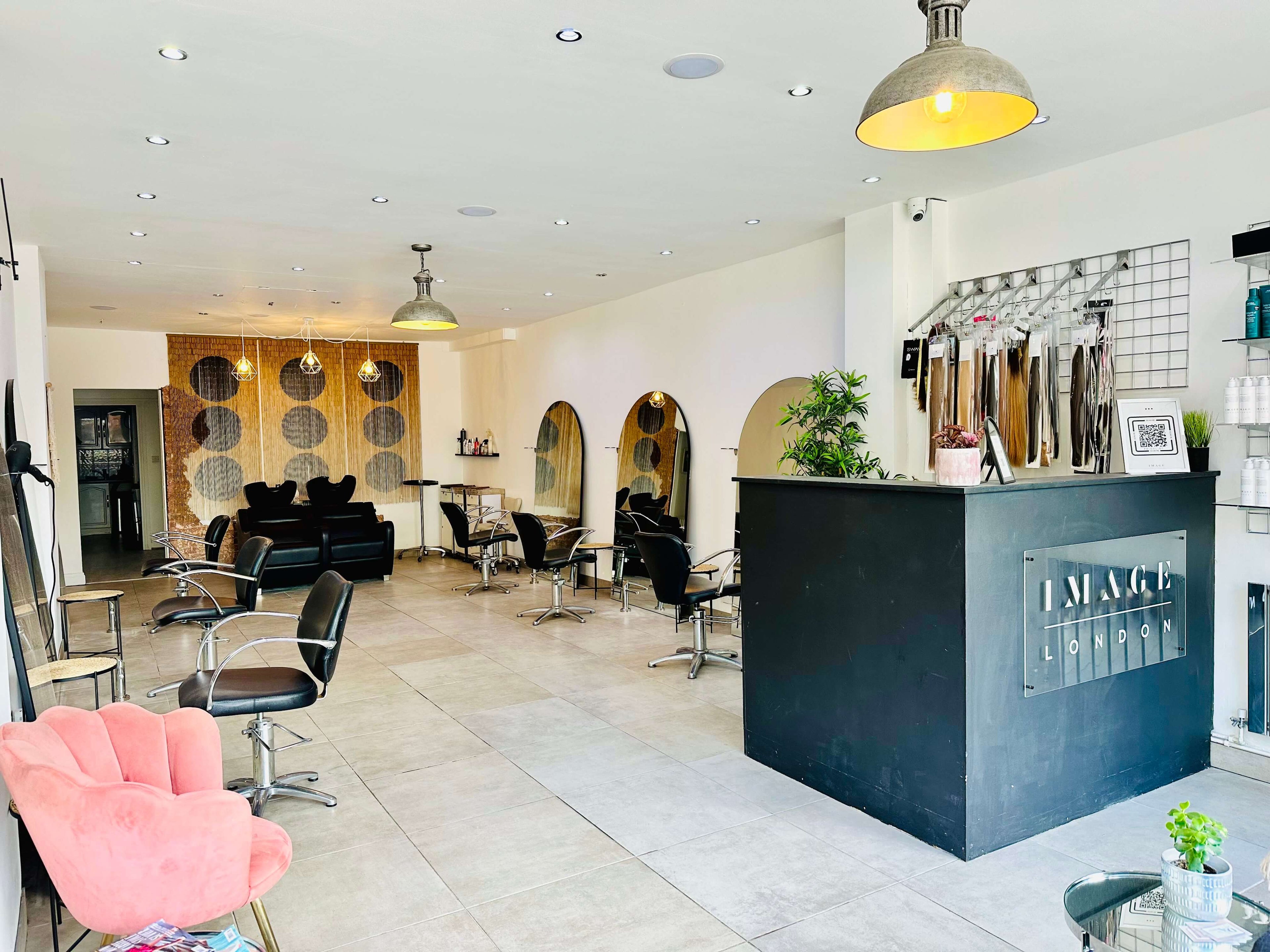 A modern, brightly lit hair salon with black styling chairs and mirrors along the left wall. The reception desk is on the right, with hair products displayed on the wall above. A pink velvet chair sits in the foreground, and large circular wall art decorates the back. We also offer Image London Hair Extension Course for those looking to master new skills.