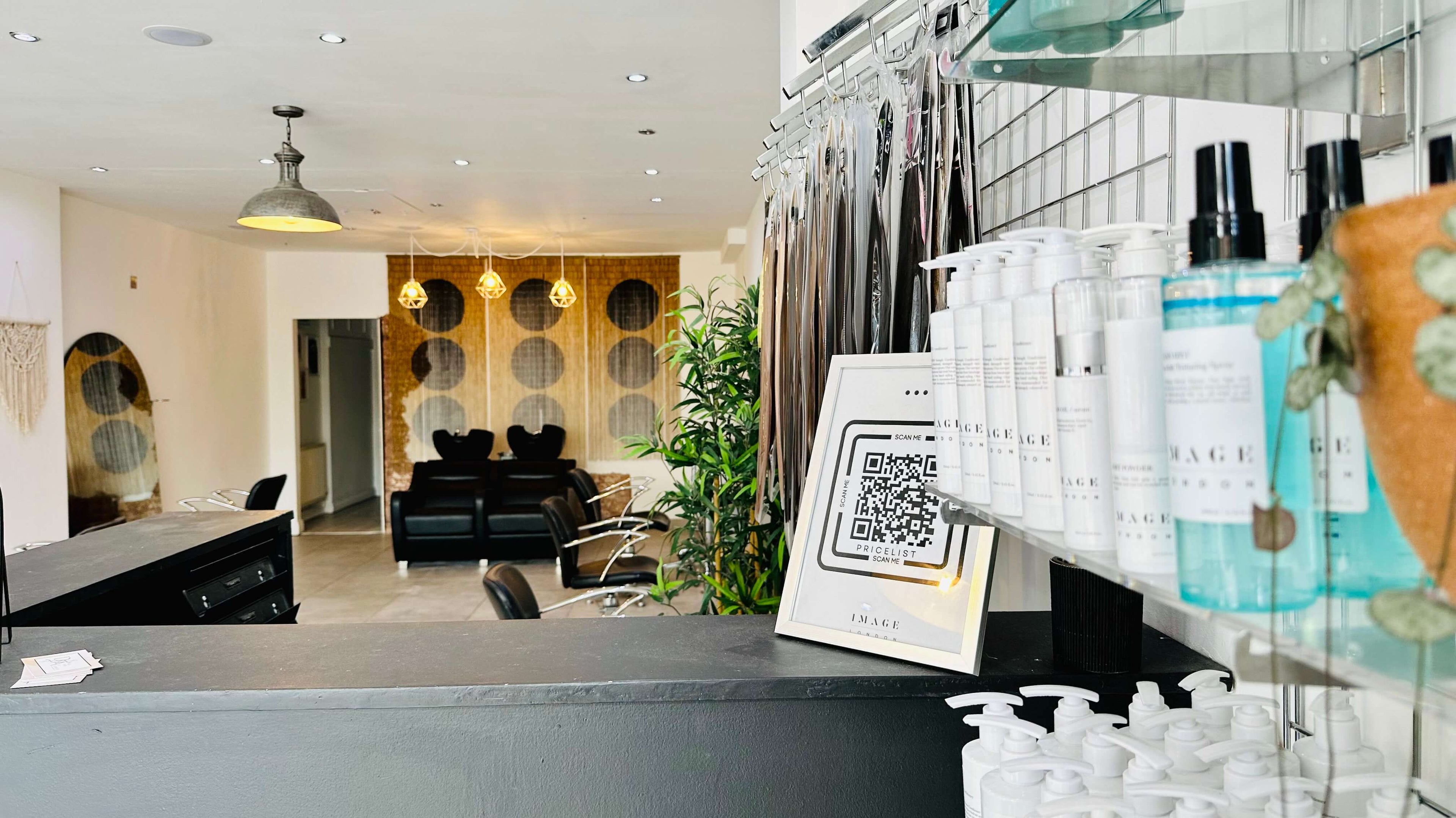 A modern hair salon with black chairs in the background and stylish decor. The foreground features a counter with Image London Products, a framed QR code, and an arrangement of hair extensions hanging on a rack. Hair extension courses are advertised, with plants and pendant lights enhancing the ambiance.