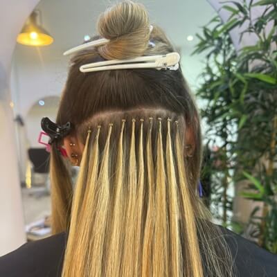 The image shows the back of a woman's head where hair extensions are being applied. Her hair is parted and held up with clips, revealing the base where blonde extensions are attached with small, noticeable loops. The setting appears to be a salon offering professional Nano Ring Hair Extension Course by Image London.